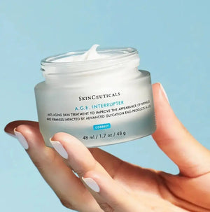 Hand holding Skinceuticals AGE Interrupter cream jar with white cream texture against blue background