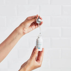 Hands holding Skinceuticals Discoloration Defense Serum bottle with dropper against white tiled background